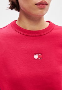 Sweatshirt rouge en tissu texturé avec un col côtelé. Présente un patch logo carré avec le texte "TOMMY" et "JEANS".