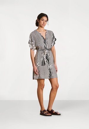 Femme debout portant une robe mi-longue à motifs noirs et blancs avec une taille nouée et des sandales marron foncé à boucle sur un fond clair uni.