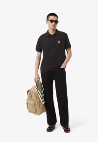 Čierna polokošeľa s logom, čierne nohavice, hnedé topánky a slnečné okuliare; drží béžový batoh so zeleným šatkou s vzorom.