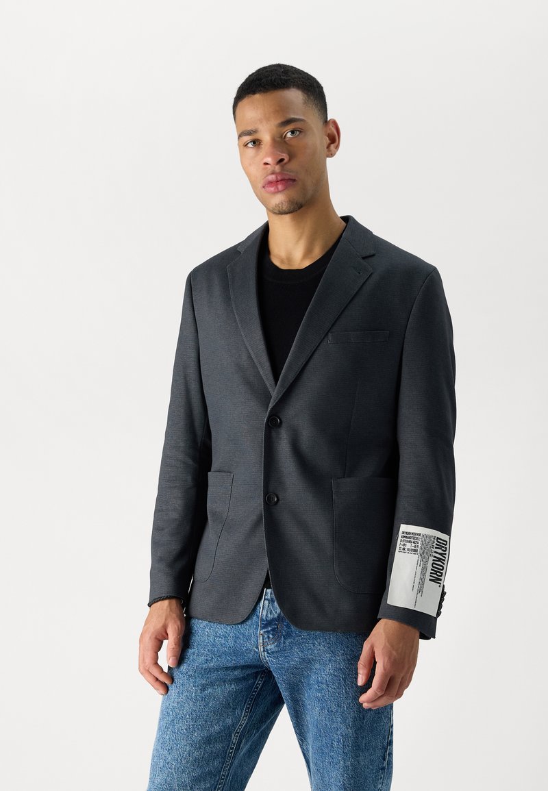Blazer gris oscuro con solapa de muesca, dos bolsillos delanteros y un detalle de parche bordado en la manga. Llevado sobre una camisa negra y unos vaqueros.
