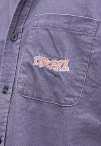 Lila kordbársony ing gombos elejű és finom textúrájú. Bal mellrészen zseb, amelyen narancssárga és kék hímzett logó található.