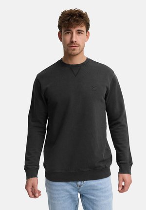 Giovane uomo con capelli corti e ricci, indossa una felpa nera a maniche lunghe e jeans azzurri, in piedi contro uno sfondo chiaro e uniforme.