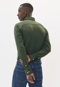 Tamnozelena košulja s dugim rukavima i šiljastim ovratnikom, s dugmićima na manžetama i teksturiranim materijalom, nosi se uz plave traperice.