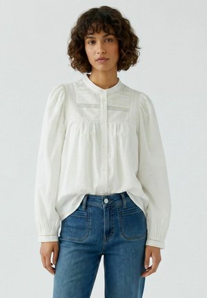 Vrouw met krullend haar, draagend een wit gebuttond blouse met pofmouwen en blauwe hooggetaillede spijkerbroek tegen een effen achtergrond.