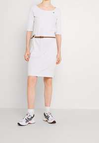 Jasnoszara bawełniana sukienka z rękawami o długości trzech kwart, dekoltem w serek i plecionym brązowym paskiem w talii. Noszona z białymi butami sportowymi.