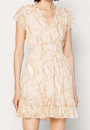 Femme portant une robe courte beige et blanche à imprimé serpent, col en V, manches flottantes, taille cintrée et ourlet volanté.