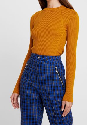 Geribbelde mosterdgele gebreide trui met een ronde halslijn, gecombineerd met blauwe geruite broek met ritsen en een aansluitende taille.