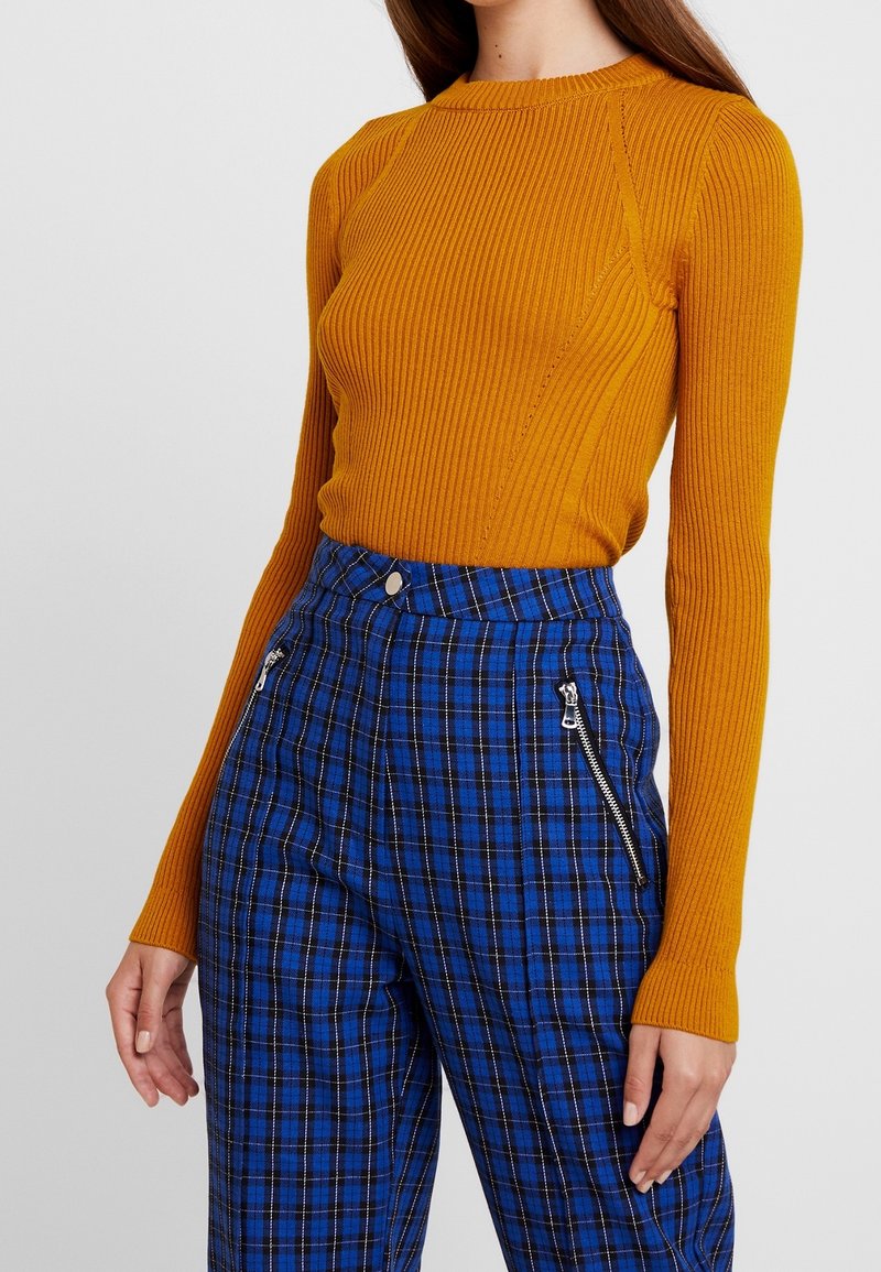 Ribbade senapsgula stickade tröja med rund halsringning, tillsammans med blå rutiga byxor med dragkedjor och figurnära midja.