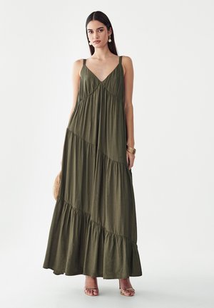 Mujer con un vestido largo sin mangas de color verde oliva con tirantes finos y costuras diagonales superpuestas, combinado con sandalias doradas y pendientes de perlas.