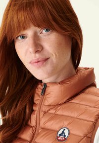 Gilet imbottito con cappuccio di colore marrone chiaro, con texture trapuntata e logo ricamato sul lato sinistro del petto. Chiusura anteriore con zip e colletto alto.