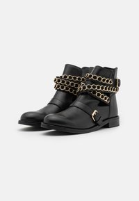 Botas de tobillo de cuero negro con correas de cadena doradas y un broche lateral. Textura suave con punta redondeada y tacón bajo.