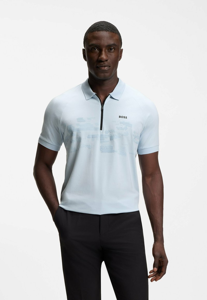 Homme portant un polo bleu clair à manches courtes avec une fermeture éclair noire et le logo "BOSS", associé à un pantalon noir, debout avec une main dans la poche.