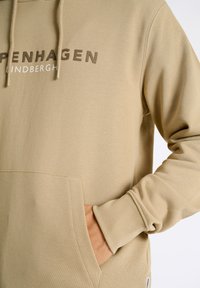 Felpa beige con cappuccio, dotata di una tasca frontale e di un logo stampato in marrone scuro. Texture liscia e polsini a coste.