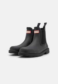 Black rubber Chelsea boots with elastic side panels, pull tabs, and a textured outsole. Features a red and white Hunter logo on the front.