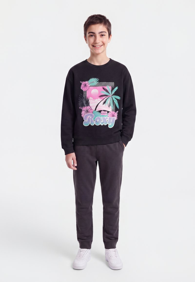 Adolescent garçon souriant, portant un sweat-shirt noir avec un motif tropical rose et bleu et un pantalon noir, debout devant un fond blanc uni.