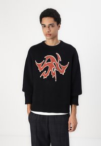 Sort strikket sweater med et stort rødt abstrakt design på forsiden, der har ribbet halsudskæring og manchetter. Bæret over en hvid skjorte.