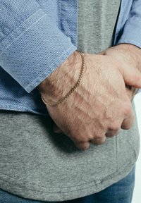 Aranyszínű fém lánc karkötő, amely egy csuklón van viselve, texturált mintával. A kéz részben látható, kék és szürke ing háttérben.