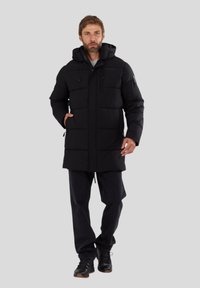 Schwarze gepolsterte Winterjacke mit Kapuze, ausgestattet mit Fronttaschen, verstellbaren Kordeln und einem Reißverschluss. Tragend mit dunklen Hosen und Schuhen.