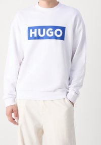 Biała bluza z niebieskim logo „HUGO”. Posiada okrągły dekolt, prążkowane mankiety i luźny krój. Miękka tkanina o gładkiej strukturze.