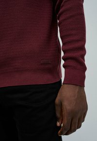 Homme portant un pull texturé marron foncé et un pantalon noir, montrant le bras droit et la main reposant à ses côtés.