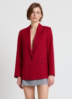 Femme portant un blazer rouge oversize avec un bouton noir et une mini-jupe grise, debout devant un fond uni gris clair.