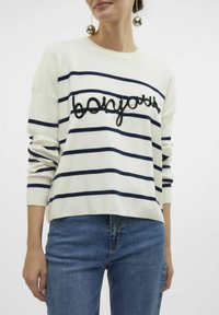 Pull en tricot blanc avec des rayures navy et le texte brodé "bonjour". Il présente une coupe décontractée et des manches longues. Associé à un jean bleu.