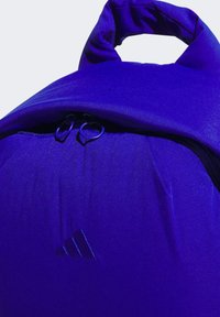 Primer plano de una mochila azul brillante con un asa acolchada, dos tiradores de cremallera y un sutil logotipo triangular en el bolsillo frontal.