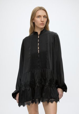 Donna bionda che indossa un vestito nero a maniche lunghe con collo alto, dettagli con bottoni, pieghe e bordatura in pizzo sull'orlo.