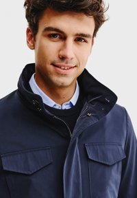 Granatowa kurtka z wysokim kołnierzem i podwójnymi kieszeniami na piersi, wykonana z gładkiego materiału. Posiada zamek błyskawiczny oraz nałożoną koszulę pod spodem.