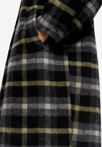 Abrigo de cuadros en negro, gris y verde con una tela suave y texturizada. Presenta un cuello de solapa y un cierre de botón en el puño.