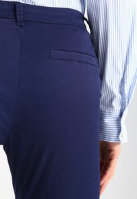 Navyblauwe broek met een soepele textuur, voorzien van een klassieke achterzak, gecombineerd met een lichtblauw gestreept shirt met knoopjes op de mouwen.