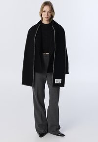 Abrigo de lana negro con costuras blancas en contraste, llevado abierto sobre una camiseta negra y pantalones grises. Presenta una etiqueta en la costura interior.