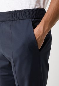 Gros plan d'une personne portant un pantalon bleu marine foncé avec une taille élastique et la main reposant dans la poche avant.