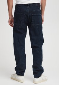 Jeans in denim blu scuro con vestibilità rilassata, caratterizzati da cuciture triple, due tasche posteriori e un passante per cintura visibile. Combinati con sneakers bianche.