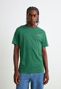 Grøn bomulds T-shirt med korte ærmer, rund halsudskæring, prydet med et lille hvidt Columbia-logo på venstre bryst. Båret med lyseblå jeans.