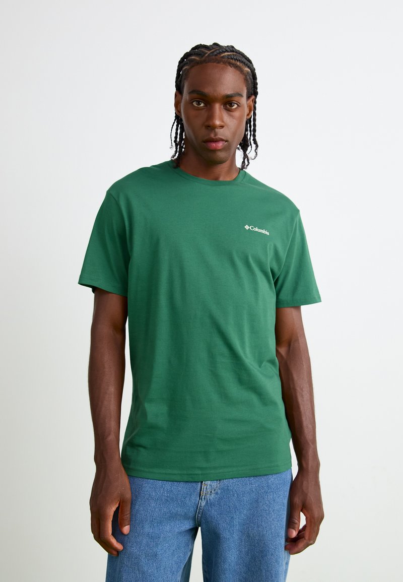 T-shirt di cotone verde con maniche corte, scollatura tonda, con un piccolo logo Columbia bianco sul lato sinistro del petto. Indossato con jeans azzurri chiari.
