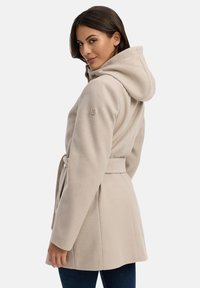Cappotto beige con cappuccio e cintura, realizzato in tessuto morbido. Presenta un dettaglio con logo sulla manica e una vestibilità su misura.