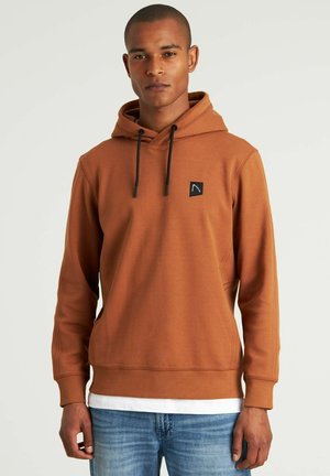 Brauner Hoodie aus weichem Stoff, mit einer Fronttasche, verstellbaren Kordeln und einem kleinen schwarzen Logo-Patch auf der Brust.