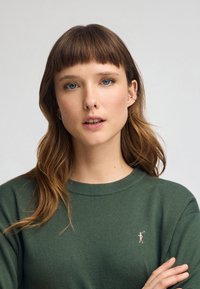 Pull en laine noir verdâtre avec col rond, arborant un petit logo rose d'un golfeur sur la poitrine, texture lisse et coupe décontractée.
