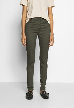 Slim fit donkerolijfgroene broek gedragen met een beige korte mouw top en zwarte instap loafers met grote gouden gespen.