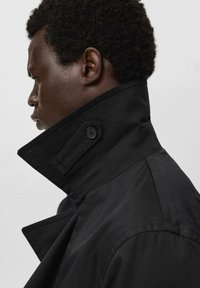 Abrigo negro con cuello alto, que presenta un detalle de botón y un diseño estructurado. La tela parece suave con textura mínima.
