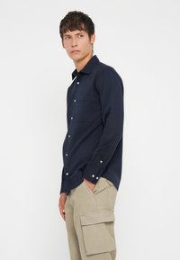 Tumši zilas krāsas krekls ar garām piedurknēm un priekšējo kabatu, izgreznots ar kontrastējošām baltām pogām un gludu auduma faktūru.