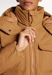 Personne fermant la fermeture éclair d'une veste matelassée beige avec de grandes poches et un col haut.