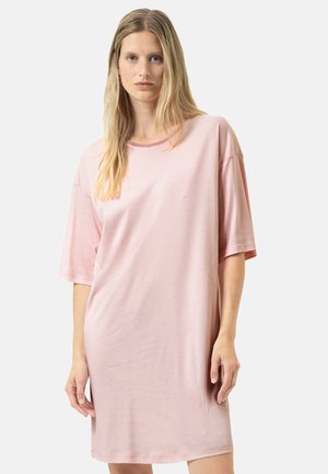 Lyshåret kvinde iført en løs, kortærmet, pink og hvidstribet kjole, der står mod en ensfarvet lys baggrund.