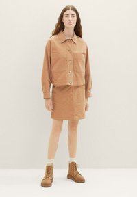 Veste en velours côtelé marron avec deux poches avant, associée à une mini jupe assortie. Portée avec des bottes beiges à lacets et des chaussettes crème.