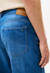 Blue denim jeans featuring a leather label with embossed text, a back pocket with a small red, white, and blue logo detail.