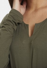 Oliwkowa bluzka z dekoltem w serek, długimi rękawami i lekko rozciągliwym materiałem. Widoczne detale szwów w okolicy dekoltu.