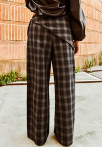 Oseba v rjavi usnjeni jakni in širokih hlačah z visokim pasom v črno-sivem karo vzorcu stoji na betonu blizu opečnate stene.