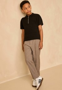 Black short-sleeve polo with a zippered collar, paired with brown plaid trousers and white sneakers with black stripes. Smooth, textured fabric.