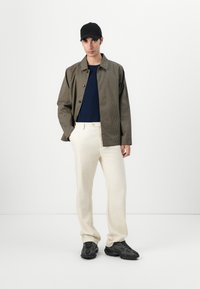 Checkered brown and black jacket, navy blue shirt, beige pants, and black sneakers, standing against a plain white background.